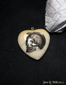 Skull scrimshaw on ivory heart with left half of the ink wiped away, the right half still covered by initial inking.