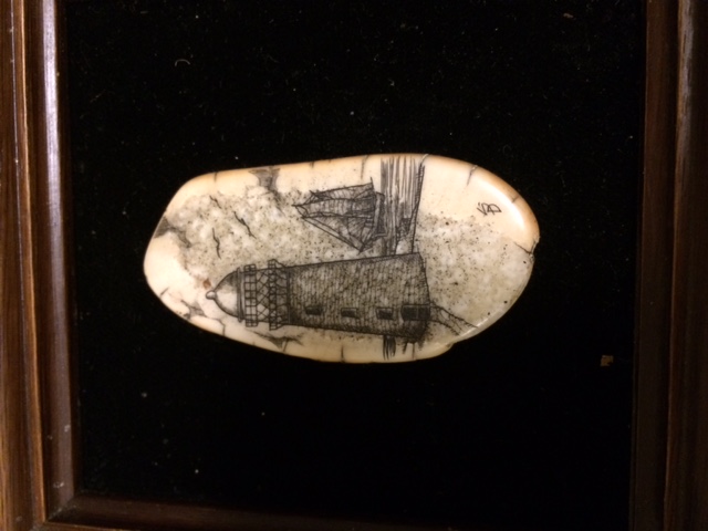 Flat tooth or bone with a domed lighthouse and a ship in the distance scrimshawed and filled with black pigment.