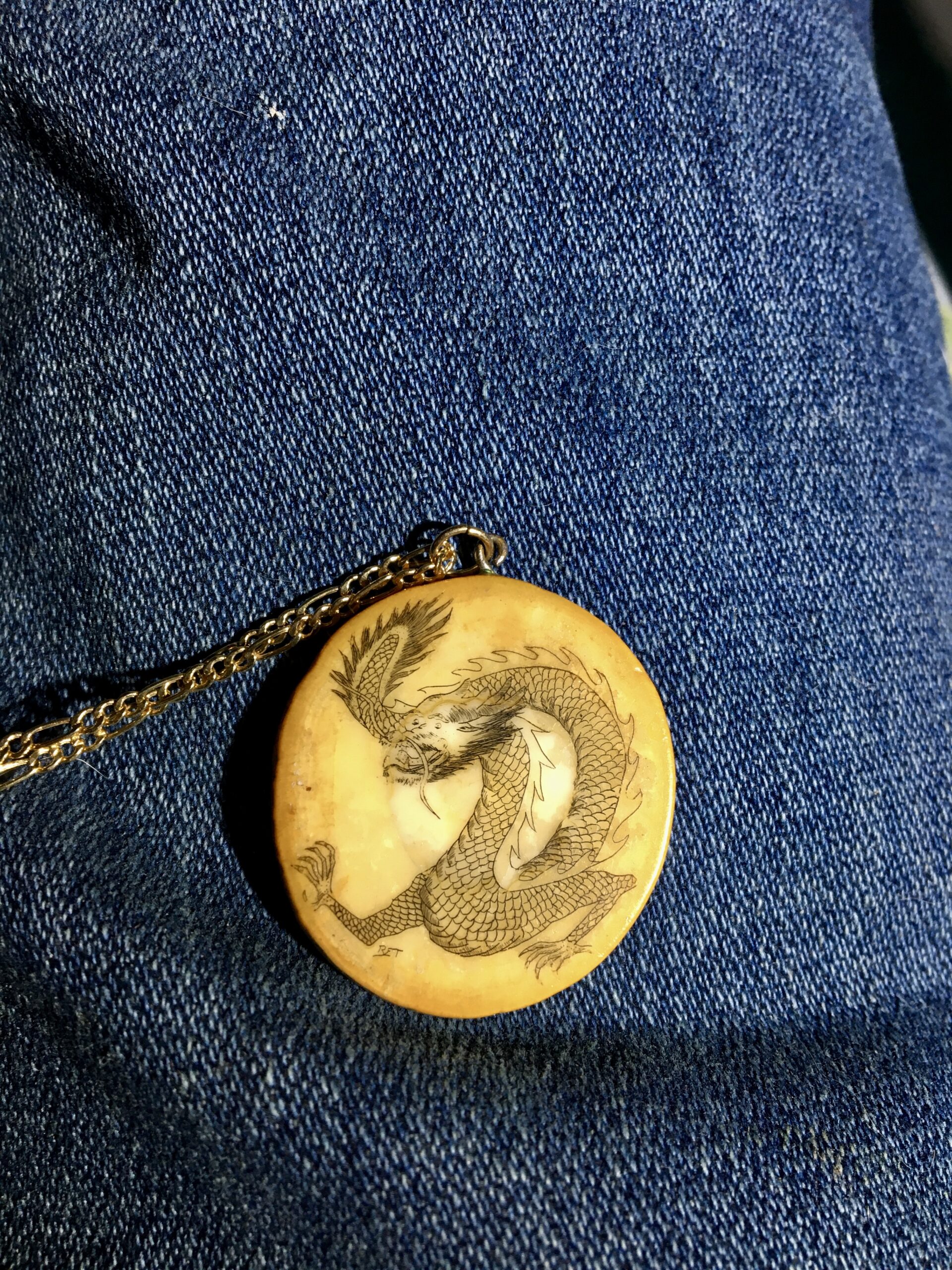 Scrimshaw of a dragon on an ivory disk with a chain attached to the top, photographed on denim